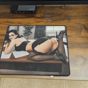 May include: A rectangular desk mat featuring a photographic print of a person with dark hair and glasses, wearing a black top, skirt, and sheer black stockings, positioned on a wooden office desk with papers scattered across the surface.