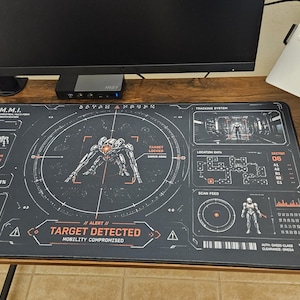 May include: A rectangular black desk mat featuring a detailed sci-fi interface design. The graphic includes a central E.M.M.I. robot, target reticles, system status text, and the bold red warning text "TARGET DETECTED" printed on the surface.