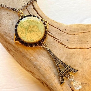 May include: A vintage-style necklace featuring a pendant with a map design and a small Eiffel Tower charm. The pendant has a gold-colored frame and a clear crystal bead. The necklace is on a brown chain.