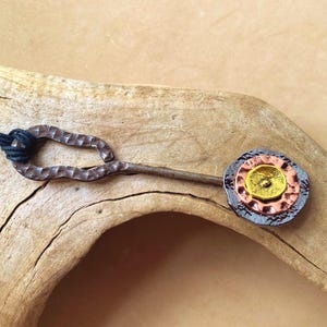 May include: Handmade metal bottle stopper with a hammered finish. The stopper features a decorative end with layers of gold, copper, and silver-colored metal. A black cord is attached to the handle. The stopper is displayed on a piece of driftwood.