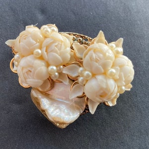 May include: A vintage floral brooch featuring ivory-colored carved flowers and faux pearls. The brooch has a gold-tone metal frame and a large, iridescent shell-shaped base. The design is intricate and detailed, with a classic, elegant aesthetic.