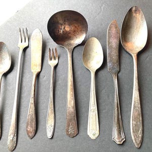 May include: A collection of seven vintage silver-plated utensils including two spoons, two forks, two knives, and one large soup ladle. Each piece features a tarnished metallic finish and decorative embossed floral patterns on the handles.