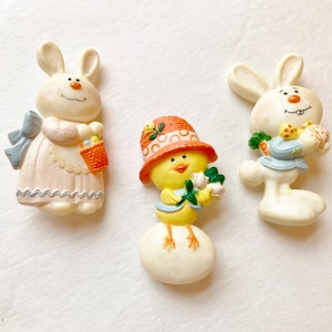 May include: Three Easter-themed figurines: two white bunnies and a yellow chick. The bunnies hold baskets and eggs, while the chick wears a hat and holds flowers. The figurines are painted in pastel colors.