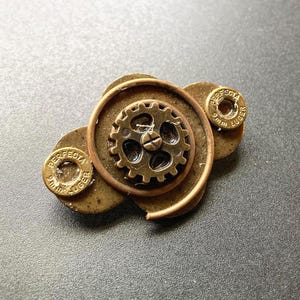 May include: A steampunk-style brooch featuring a small gear at the center, surrounded by a spiral of metal and two bullet casings. The brooch has a bronze and copper color scheme, with the text "9mm LUGER" visible on the casings.