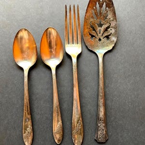 May include: A set of four silver-toned metal utensils including two spoons, one four-tined fork, and one wide, perforated cake server. Each piece features a tarnished finish and an embossed floral pattern along the handle stems.