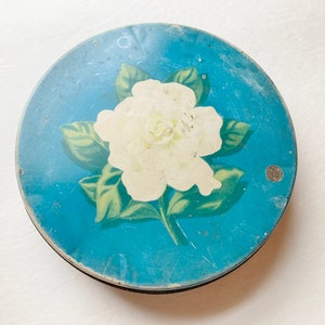 May include: A round, vintage tin with a light blue background. A white flower with green leaves is centered on the lid. The tin has a black rim and shows signs of wear.