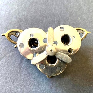 May include: A decorative brooch featuring three silver-toned metal sewing machine bobbins arranged in a triangular cluster. A small, three-bladed silver propeller is centered over the bobbins, and two ornate, antiqued bronze-colored metal wings extend from the sides.