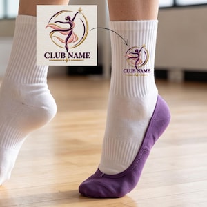 Op de afbeelding: Een paar witte sokken met een paars en goudkleurige afbeelding van een dansend figuur in een cirkelvormig kader met de tekst CLUB NAME eronder. De sokken worden gedragen in combinatie met paarse, laag uitgesneden sokken.