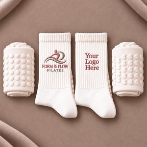 Puede incluir: Dos calcetines de corte medio acanalados en color blanco roto y dos calcetines enrollados con agarres de silicona. Un calcetín tiene un logo bordado con una figura estilizada y el texto FORM & FLOW PILATES. El otro tiene el texto Your Logo Here.