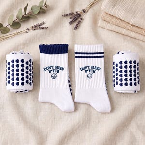 Puede incluir: Dos calcetines blancos con puños acanalados y rayas azul oscuro, con el texto DON'T SLEEP B*TCH y un icono de cara durmiente. Otros dos calcetines blancos están enrollados, mostrando un patrón repetitivo de pequeños corazones azul oscuro.