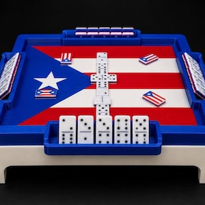 May include: A square domino game table featuring a blue frame and white base. The playing surface displays the Puerto Rican flag with a blue triangle, white star, and red and white stripes. White dominoes with black pips are arranged on the board.