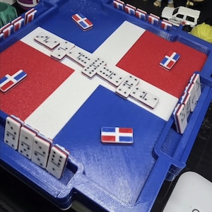 May include: A blue, white, and red domino game board featuring the Dominican Republic flag design. The board includes integrated slots for standing domino tiles, with several tiles placed on the surface and in the slots, each decorated with the same flag pattern.