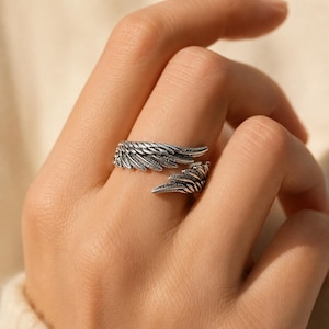 May include: A silver-toned open ring featuring a detailed wing design that wraps around the finger. The metal has an oxidized finish in the recessed areas to highlight the textured feather patterns along the length of the wings.