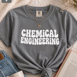 May include: A grey short-sleeved t-shirt featuring the white text "CHEMICAL ENGINEERING" in a wavy, retro-inspired font. The shirt has a crew neckline and a Comfort Colors brand label at the inner collar. A gold-toned bow necklace rests on the chest.