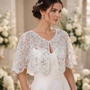May include: A sheer white bolero capelet featuring intricate silver sequin and bead embroidery in a scalloped fan pattern. The garment has an open front design with short sleeves and a scalloped hem, worn over a plain white sleeveless dress.