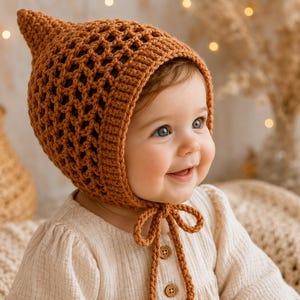 May include: A rust-colored crochet bonnet featuring a pointed top, an open lattice stitch pattern, and a ribbed border around the face. The bonnet includes two long, matching crochet ties that fasten under the chin, creating a secure fit.