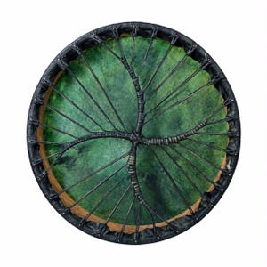 Può includere: Un tamburo a cornice circolare con una pelle di colore verde brillante dalla texture screziata. La pelle è fissata a un telaio in legno tramite un sistema di allacciatura nero che converge al centro, creando un motivo a croce sulla superficie.