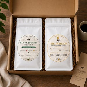 Puede incluir: Una caja de regalo de cartón que contiene dos bolsas de café blancas. La bolsa izquierda está etiquetada como Honey Journey con detalles verdes, y la derecha como The Horizon con detalles dorados. Incluye una pequeña tarjeta de marca.