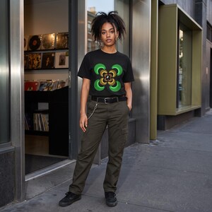 Könnte beinhalten: Ein schwarzes Kurzarm-T-Shirt mit einer zentralen Grafik aus vier symmetrischen grünen, geschwungenen Formen, die ein braun-beiges Spiralmuster mit einem kleinen Yin-Yang-Symbol umgeben, kombiniert mit einer olivgrünen Cargohose und einem schwarzen Gürtel.