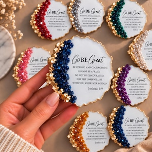 May include: A collection of round white coasters with irregular gold-trimmed edges and clusters of colored crystals. Each coaster features the black text: GO BE GREAT, BE STRONG AND COURAGEOUS; DO NOT BE AFRAID; DO NOT BE DISCOURAGED, JOSHUA 1:9.