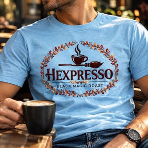 May include: A light blue t-shirt featuring a graphic of a coffee cup and a broomstick inside an oval wreath of autumn leaves. The text reads HEXPERSSO and BLACK MAGIC ROAST in a dark red font. The design is centred on the chest of the shirt.