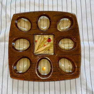 May include: A square brown wood-grain serving tray featuring eight oval-shaped indentations surrounding a central square compartment. The center square contains a decorative arrangement of dried wheat stalks, small yellow flowers, and a red ladybug.