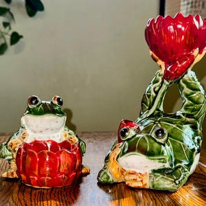 May include: Two ceramic frog figurines with a glossy finish. One frog sits upright holding a red lotus-shaped bowl. The second frog is positioned lying on its back, holding a large red lotus flower bloom upward with its hind legs and front paws.
