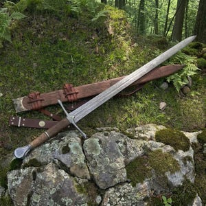 Könnte beinhalten: Ein zweischneidiges Stahlschwert mit gemusterter Klinge, Holzgriff und Metallknauf. Das Schwert liegt diagonal über einer braunen Lederscheide mit geprägten Details und Lederriemen, die mit Metallschnallen versehen sind.