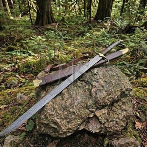 May include: A long metal sword with a dark, textured blade and a gold-toned crossguard. The sword rests diagonally across a brown leather scabbard with gold-colored metal fittings at the tip and throat, featuring an attached brown leather strap.
