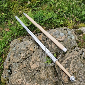 May include: A double-edged steel sword with a long, straight blade, a crossguard, and a light-colored grip, accompanied by a matching scabbard with a metal tip. The blade features a central fuller, and the hilt includes a circular pommel.