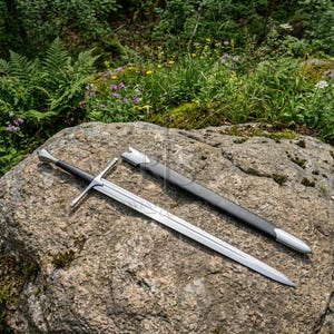May include: A long steel sword with a black grip, a straight crossguard, and a pointed blade, accompanied by a matching black scabbard with silver-toned metal fittings at the tip and throat, resting on a textured gray stone surface.