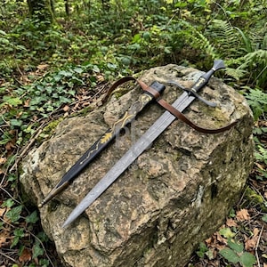 Peut inclure: Une épée à double tranchant avec une lame en acier damassé et une poignée noire ornée d'une croix dorée. Elle est accompagnée d'un fourreau noir décoré de motifs dorés en forme de feuilles et d'une sangle en cuir marron.