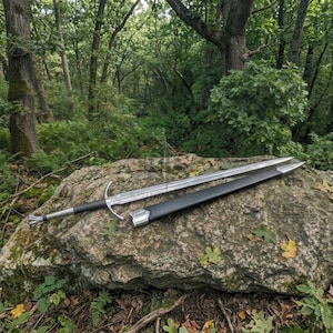 Puede incluir: Una espada de acero de doble filo con una guarda en forma de cruz y empuñadura negra, acompañada de una vaina negra a juego con herrajes de metal plateado. La espada y la vaina están colocadas horizontalmente sobre una gran roca gris.