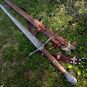 Könnte beinhalten: Ein Langschwert mit gemusterter Stahlklinge, einem Parier mit kleinem grünen Stein und einem braunen, lederumwickelten Griff. Dazu gehört eine passende braune Lederscheide mit aufwendigen Prägemustern und metallischen Akzenten.