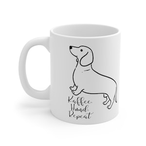 Könnte beinhalten: Ein weißer Keramikbecher mit einer schwarzen Strichzeichnung eines Dackels. Unter dem Hund steht der Text "Kaffee. Hund. Repeat" in einer Kombination aus schwarzer Schreib- und Druckschrift. Der Becher hat einen geschwungenen Henkel links.
