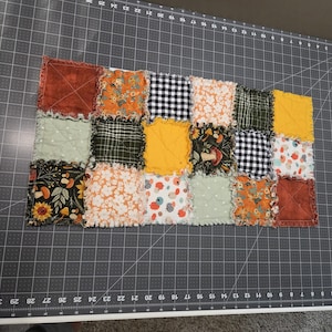 May include: A rectangular rag quilt featuring a patchwork of fabric squares in various patterns and colours, including mustard yellow, sage green, burnt orange, black and white gingham, and floral prints, with exposed frayed seams between each square.