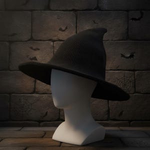 Puede incluir: Un sombrero de bruja de punto negro con una corona puntiaguda y un ala ancha y flexible. El sombrero se muestra sobre una cabeza de maniquí blanca lisa, destacando el tejido texturizado y la forma estructurada de la prenda de cabeza.