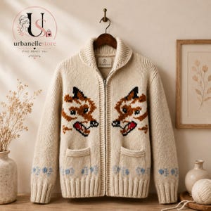 May include: A cream-colored knit zip-up cardigan featuring a shawl collar and two front pockets. The garment displays a repeating brown and black fox face pattern across the chest and a light blue paw print design along the ribbed hem.