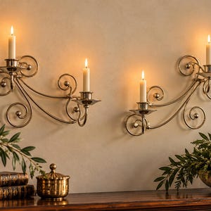 May include: Two silver-toned metal wall sconces with scrolling wire designs, each holding two lit white taper candles. The sconces feature circular accents and are mounted on a plain wall, providing a warm glow from the four burning candles.