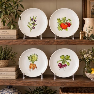 May include: Four white ceramic plates displayed on wooden shelves, each featuring a distinct botanical illustration: a branch of purple olives, a bright red tomato, a bunch of orange carrots, and a cluster of deep purple radishes with green leaves.