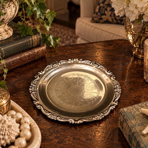 Puede incluir: Una bandeja redonda de metal plateado con un borde festoneado ornamentado con detalles en relieve y un área circular central decorada con intrincados grabados florales simétricos y un patrón de borde trenzado.
