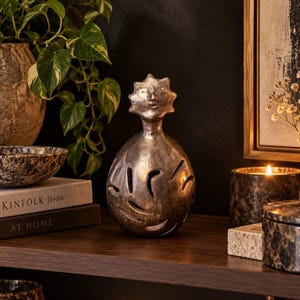 May include: A metallic silver-toned decorative sculpture featuring a rounded body with carved abstract cutouts and a stylised head with a sunburst-shaped crown. The object has a smooth, reflective finish and sits upright on a flat surface.
