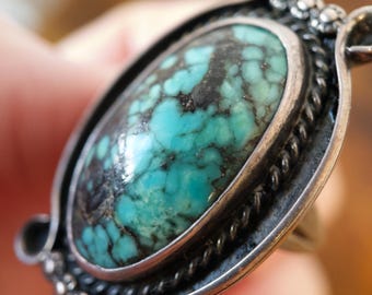 May include: A silver ring featuring a large, oval-shaped turquoise stone with dark matrix veining. The stone is set in a smooth bezel, surrounded by a twisted rope border and decorative silver accents at the top and bottom edges of the setting.