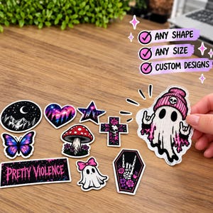 Pode incluir: Uma cole&ccedil;&atilde;o de autocolantes recortados com v&aacute;rios designs, incluindo uma lua crescente, um cora&ccedil;&atilde;o, uma estrela, uma borboleta, um cogumelo, uma cruz, um fantasma com gorro rosa, um caix&atilde;o e um autocolante retangular com o texto PRETTY VIOLENCE.