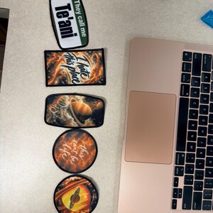 Pode incluir: Cinco emblemas de tecido com bordas pretas dispostos verticalmente ao lado de um laptop. Os designs incluem o texto "They call me Te'ani", "I hate this place", "Dreaming of Saturn", "this can't be life" e um gr&aacute;fico do planeta Saturno.