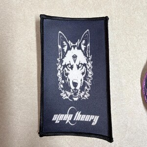 Peut inclure: Un &eacute;cusson rectangulaire en tissu noir pr&eacute;sentant un motif blanc repr&eacute;sentant une t&ecirc;te de loup avec un symbole de diamant sur le front. Sous le loup, le texte "sleep theory" est imprim&eacute; dans une police blanche stylis&eacute;e. Bordure noire cousue.