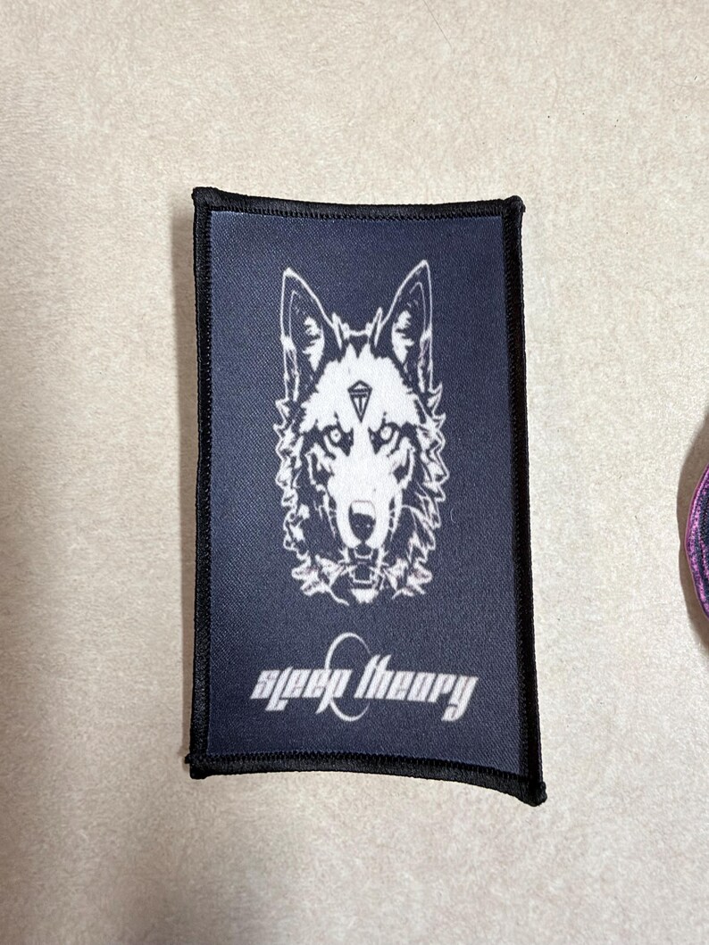 Peut inclure: Un &eacute;cusson rectangulaire en tissu noir pr&eacute;sentant un motif blanc repr&eacute;sentant une t&ecirc;te de loup avec un symbole de diamant sur le front. Sous le loup, le texte "sleep theory" est imprim&eacute; dans une police blanche stylis&eacute;e. Bordure noire cousue.