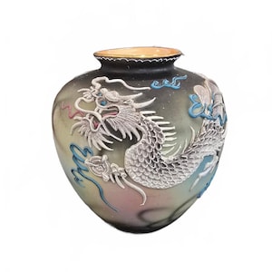 Könnte beinhalten: Eine bauchige Keramikvase mit matter, schwarz-verlaufender Oberfläche. Das Äußere ist mit einem erhabenen, strukturierten Relief eines weißen Drachen mit blauen und rosa Akzenten verziert. Der Drache windet sich um den Körper des Gefäßes.