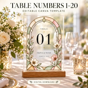 May include: A rectangular clear acrylic table number sign featuring a stained glass style arch design with floral illustrations in soft pink and green. The sign displays the text TABLE 01, OLIVIA & NOAH, and JUNE 12, 2027, set in a wooden base.