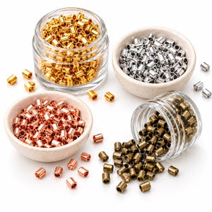 May include: A collection of small cylindrical metal crimp beads in gold, silver, copper, and antique bronze tones. The beads are organized in two clear glass jars and two small speckled ceramic bowls, with several loose beads scattered around.